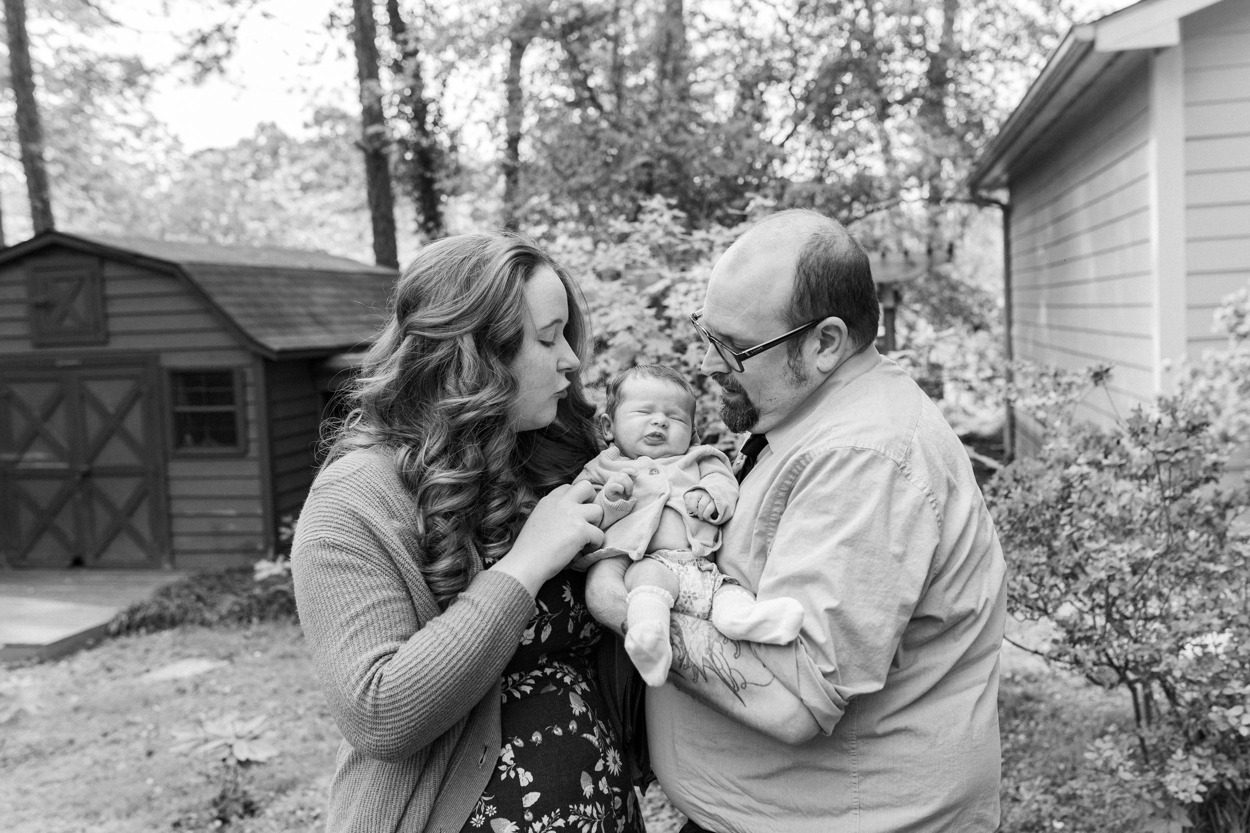 Black and white photo of parents lovingly gazing at their newborn baby outdoors near a home.