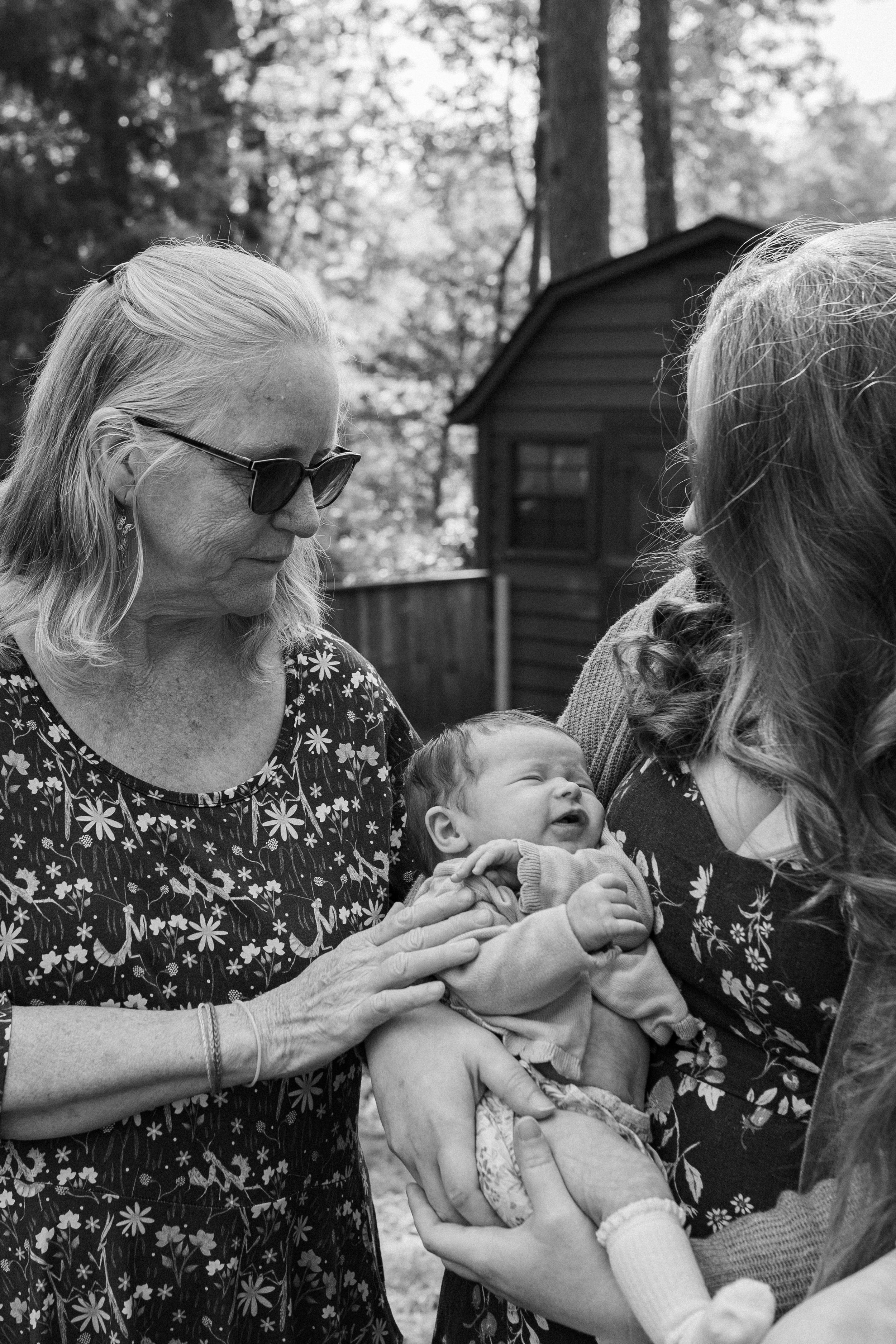 Black and white photo of grandmother and mother admiring newborn baby outdoors near wooden shed.