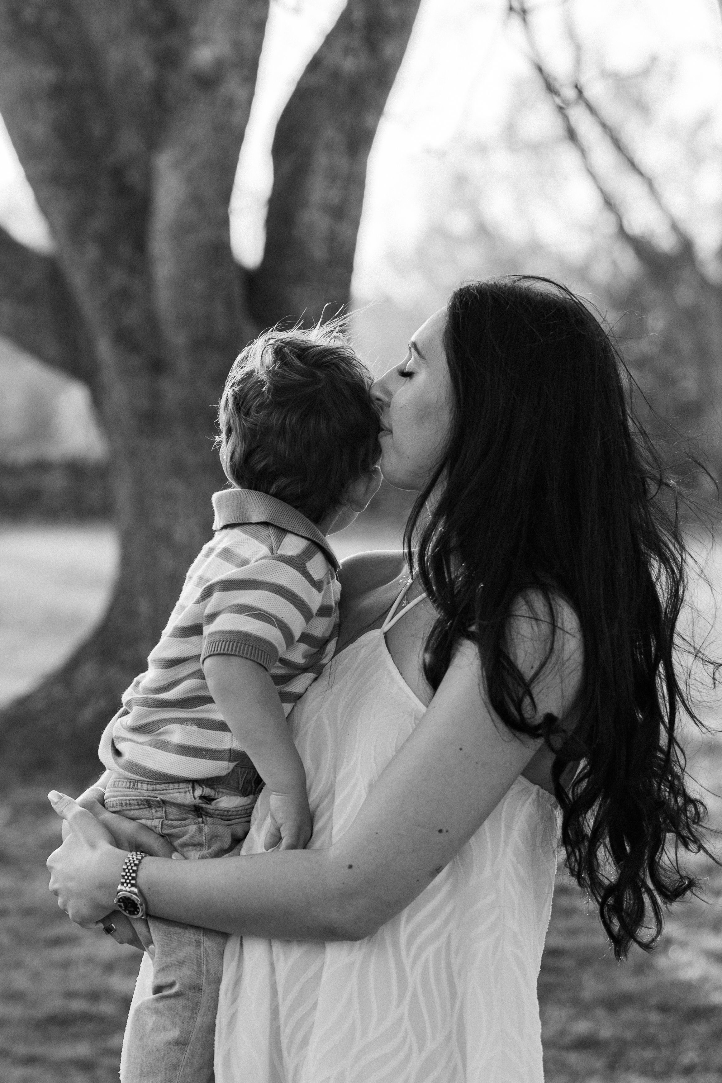 Black and white photo of a mother kissing her young son while holding him outdoors near a tree.