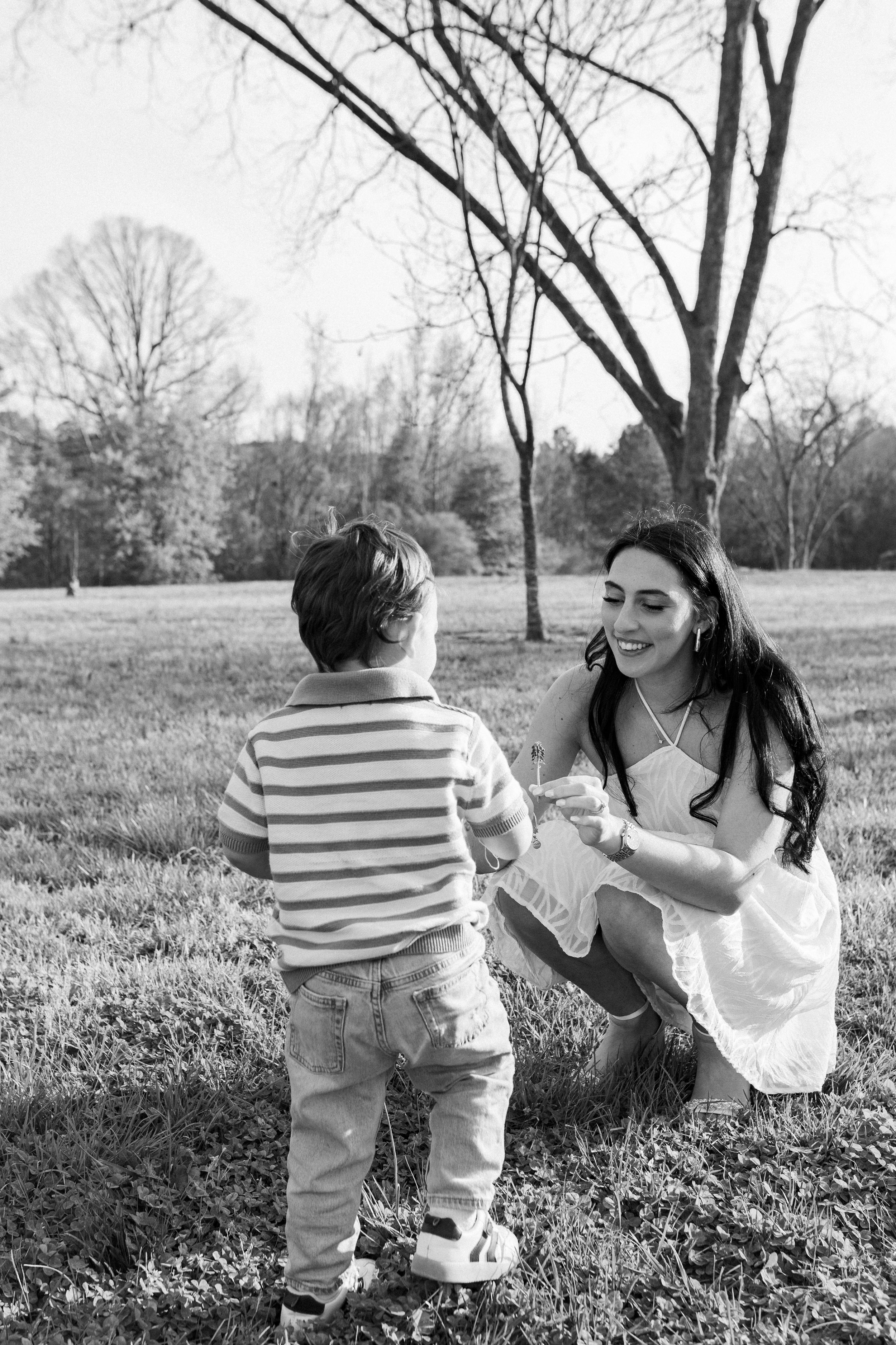 Black and white photo of a mother crouching in a park, smiling at her young toddler son standing before her.