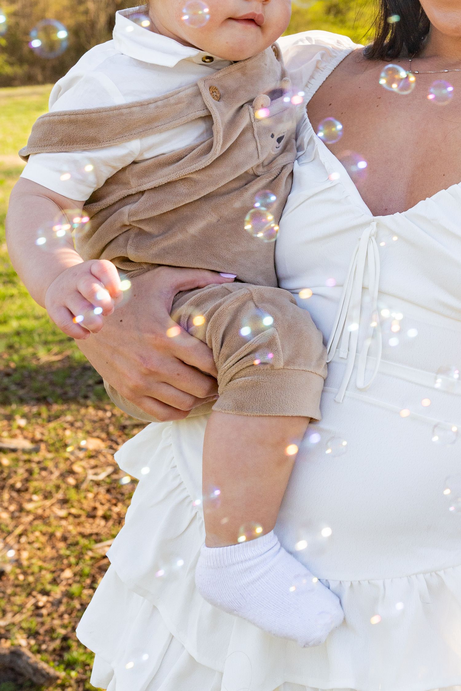 A small child wearing tan shorts and white socks sits in someone's arms while soap bubbles float through the sunlit air.