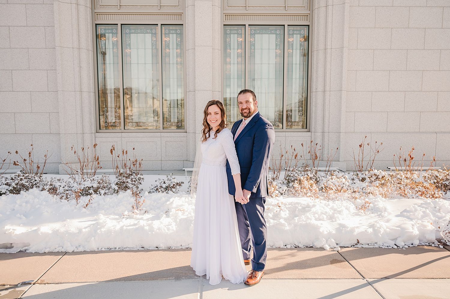 A couple stands together in winter wedding attire outside a stone building with snow on the ground.