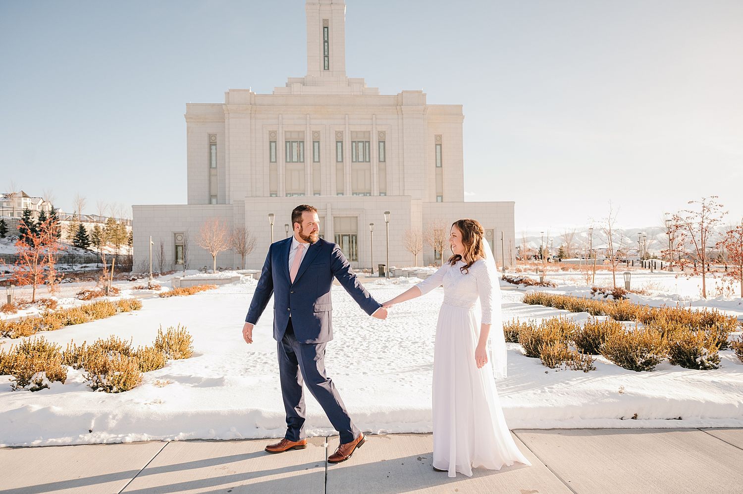 A couple in wedding attire holds hands while walking in snow in front of a white temple building.