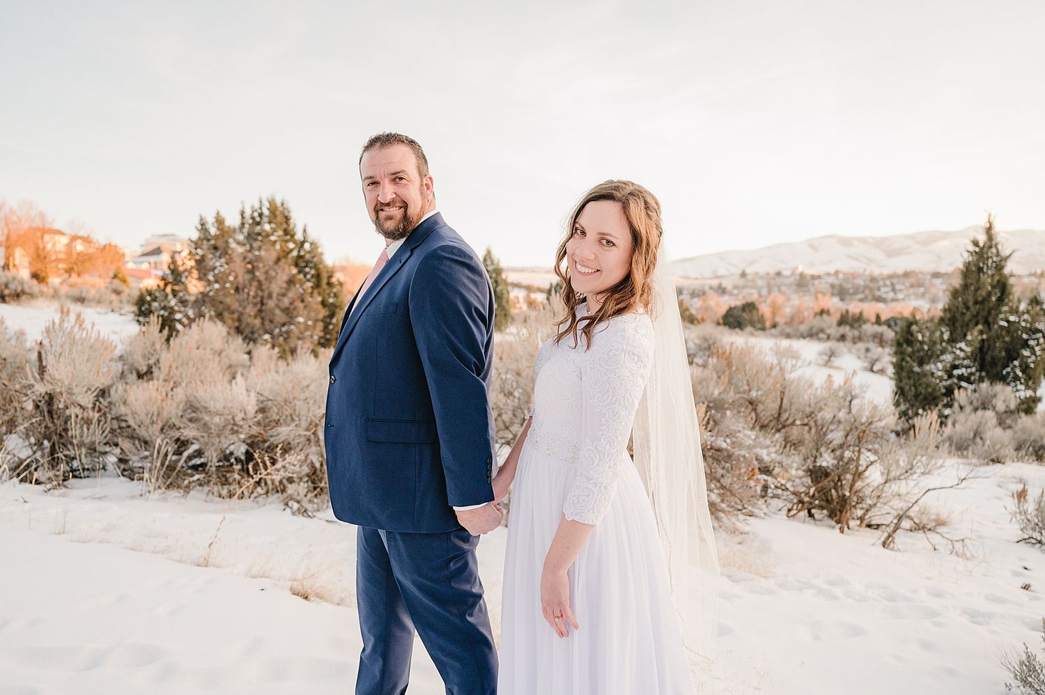A couple in formal attire standing together in a snowy desert landscape with mountains at sunset.
