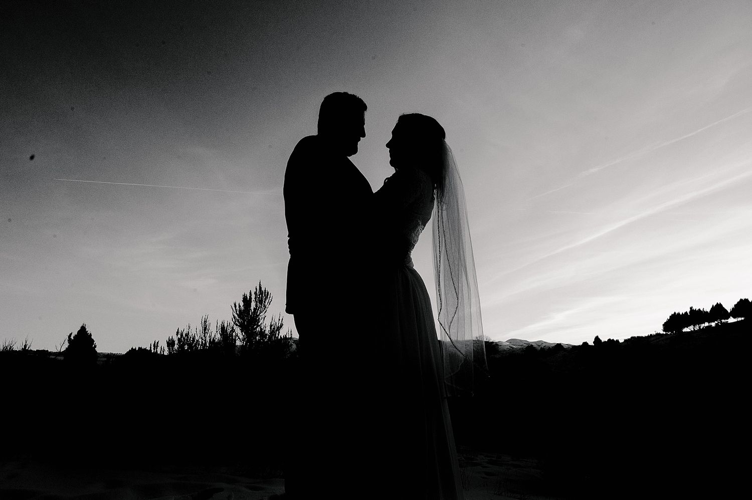 A romantic silhouette of newlyweds against a dramatic sunset sky, with the bride's veil flowing in the breeze.