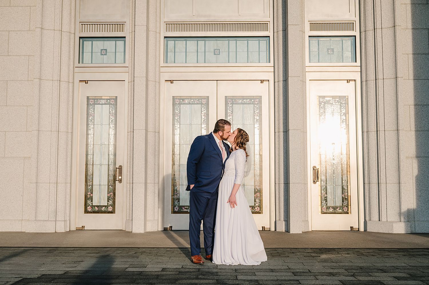 A couple in wedding attire shares a romantic moment in front of elegant white double doors at a temple entrance.