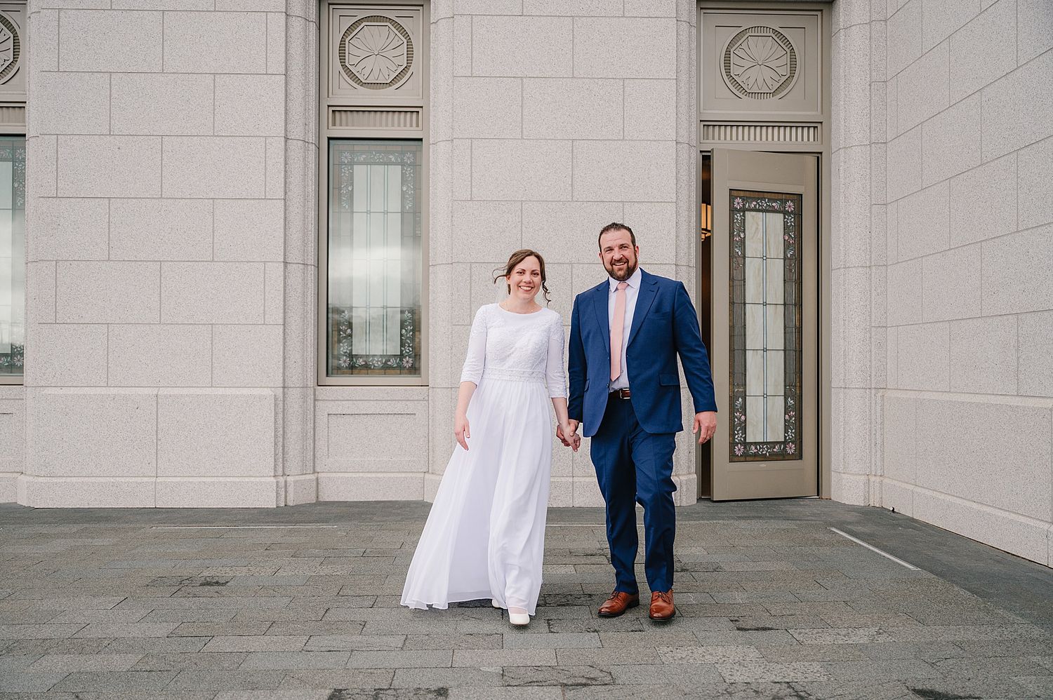 Newlyweds walking together outside a white stone building, with the bride in white and groom in navy blue.