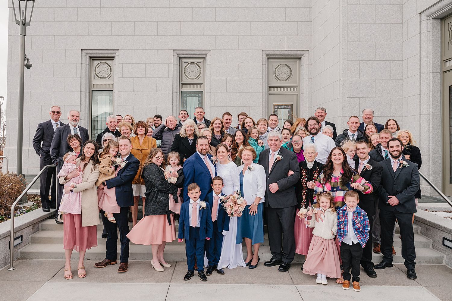 Large wedding party group photo outside a white stone building with family and friends dressed in formal attire.