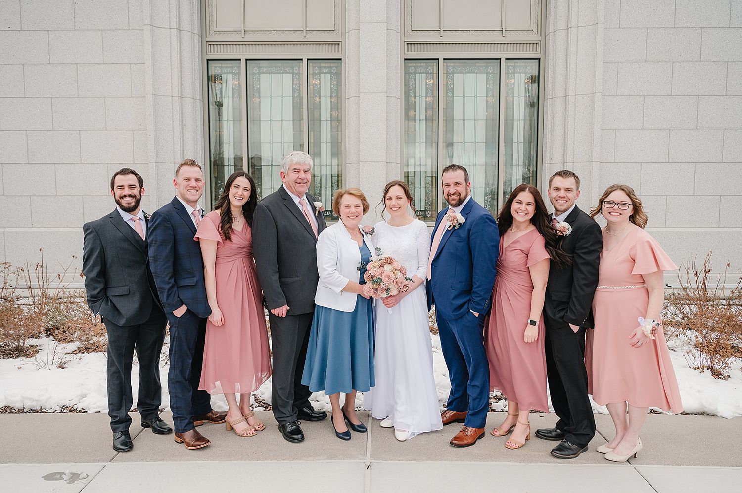 A formal group portrait of wedding party members standing together outside a white stone building in coordinated attire.