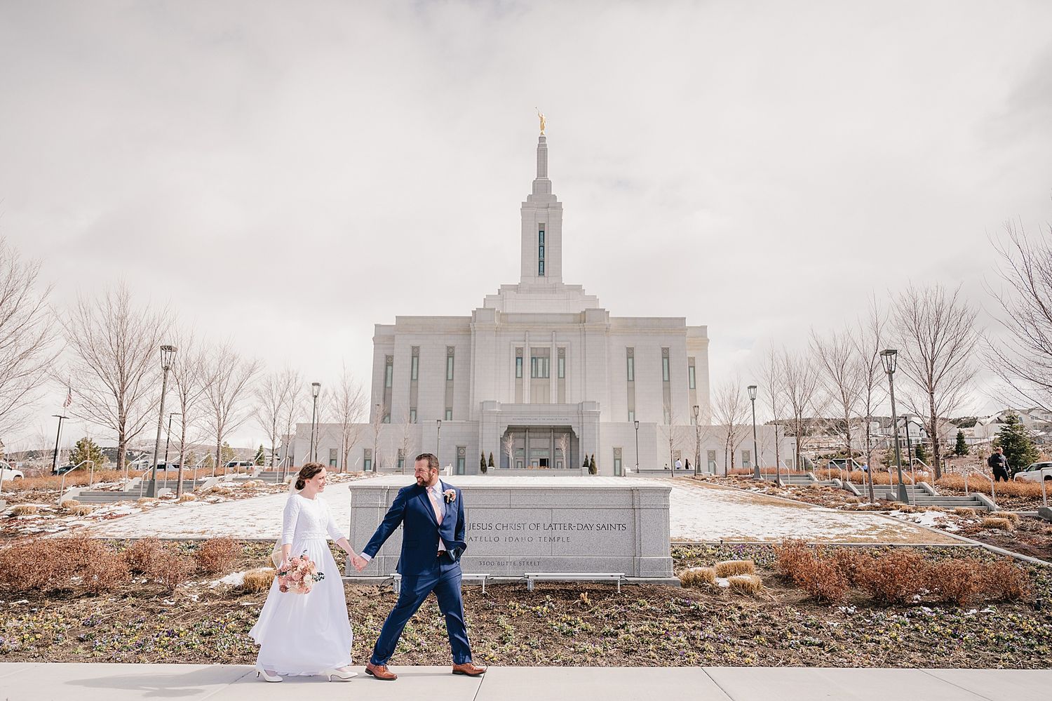 A couple in wedding attire walking hand in hand in front of the Pocatello Idaho Temple on a cloudy winter day.