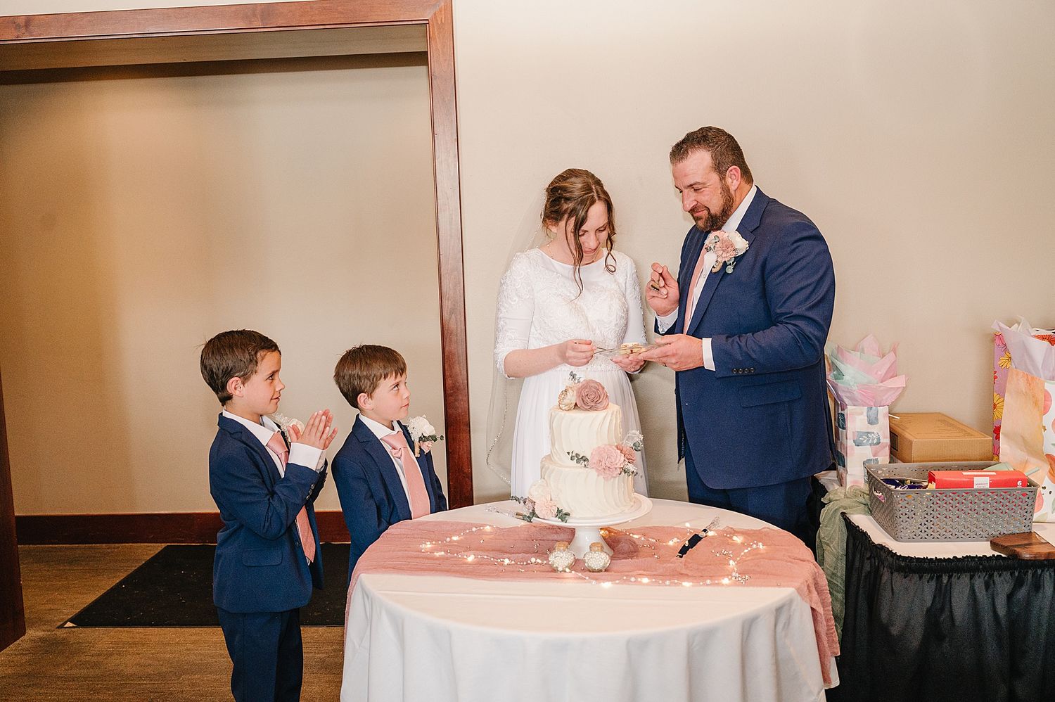 A wedding couple and two young boys in navy suits gather around a pink-decorated cake table during a celebration.