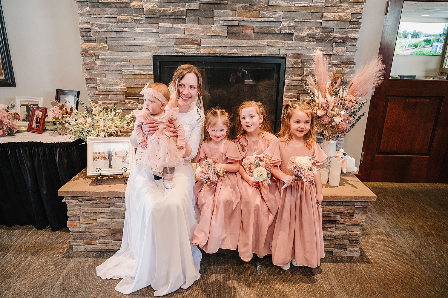 A bridal party poses in front of a stone fireplace wearing elegant white and dusty rose colored dresses.