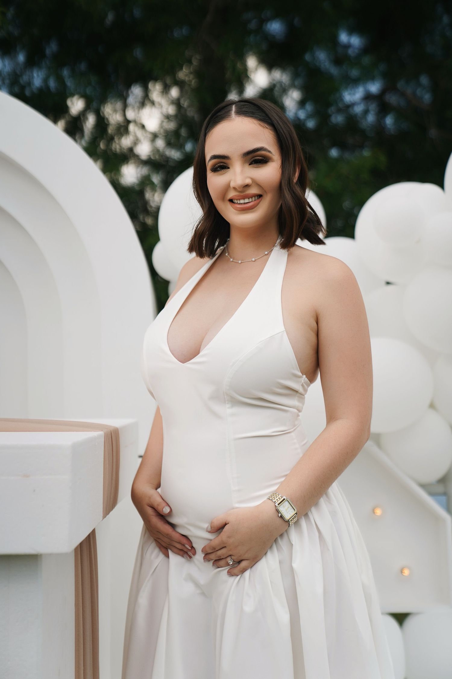 Smiling pregnant woman in white halter dress cradling baby bump at elegant white balloon baby shower.
