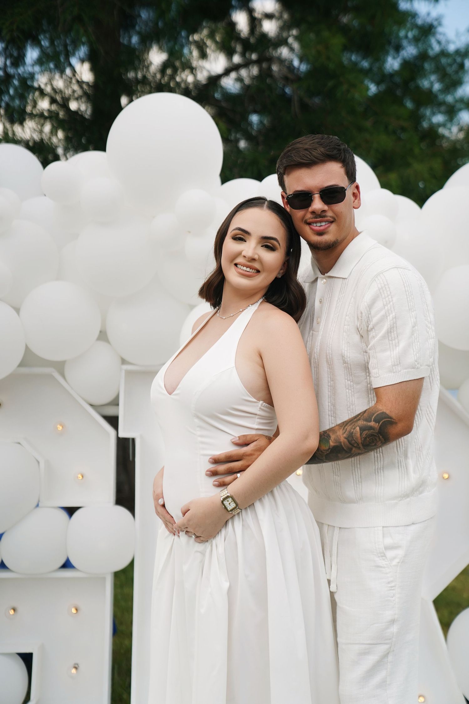 Smiling pregnant couple in white outfits pose together outdoors in front of white balloon arch decoration.
