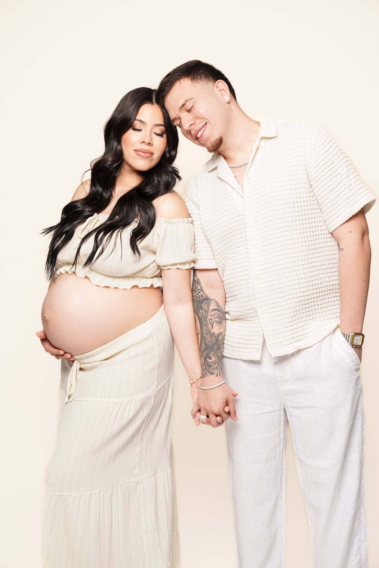 Couple in white outfits hold hands during maternity photoshoot against light background.