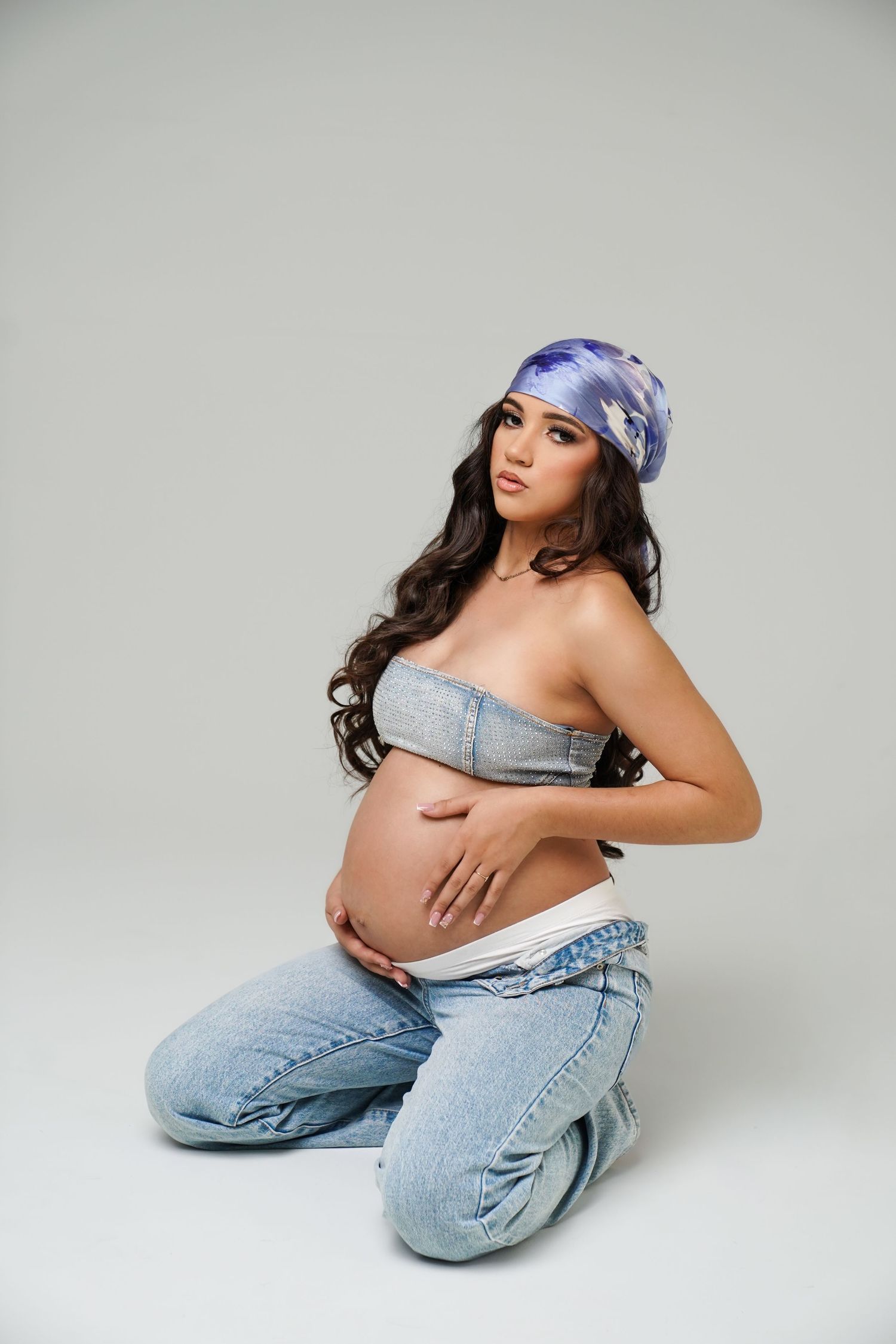 Pregnant woman in denim crop top and jeans, kneeling while cradling baby bump, wearing purple headscarf on gray background.