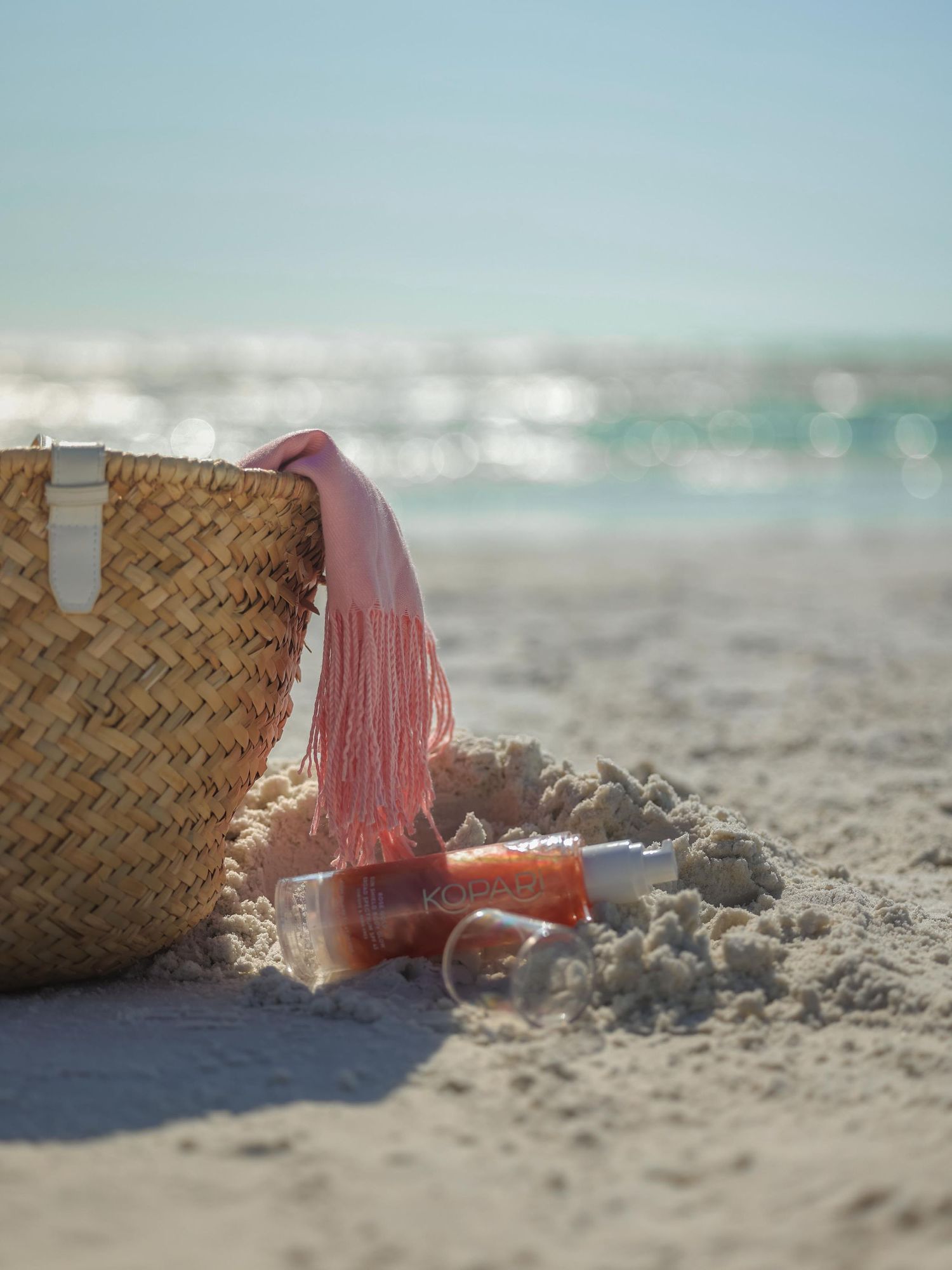 Straw beach bag and pink accessories resting on sandy shore with sparkling ocean waves in the background.