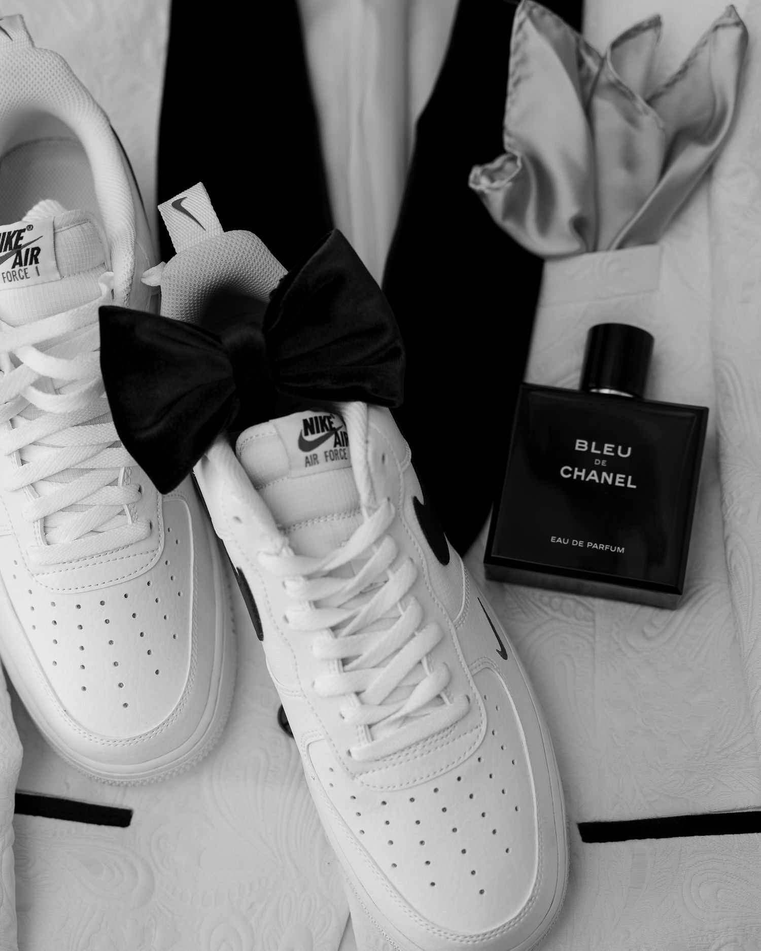 A luxury lifestyle photo featuring white Nike Air Force 1 sneakers with a black bow tie and Bleu de Chanel perfume.