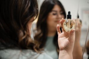A person holds up a clear glass dropper bottle of beauty serum or essential oil in front of a mirror.