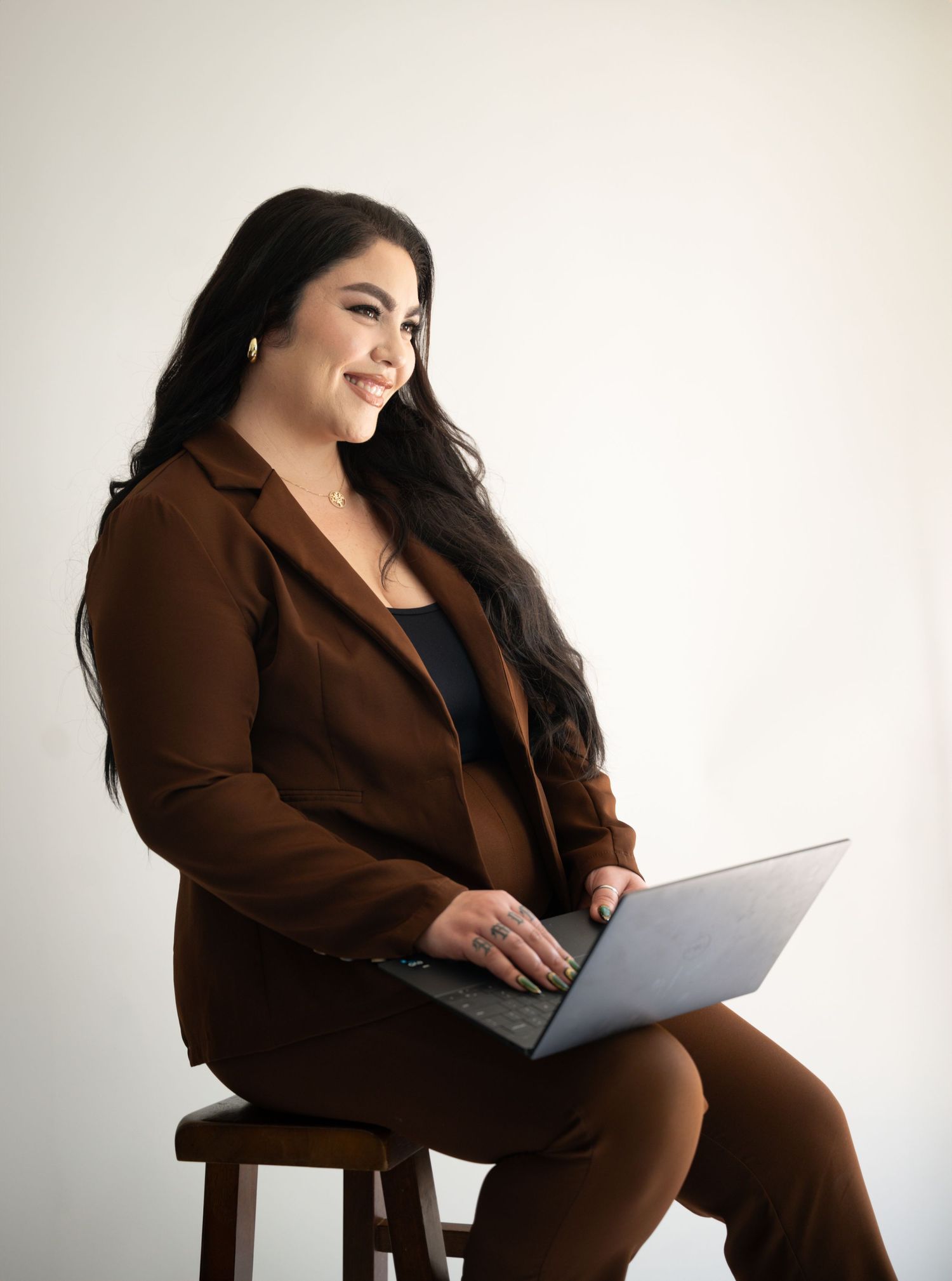 Professional wearing a brown blazer sits on a stool with a laptop against a light background.