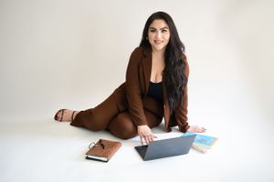 Professional in brown suit works on laptop while sitting on floor with books and papers in studio setting.