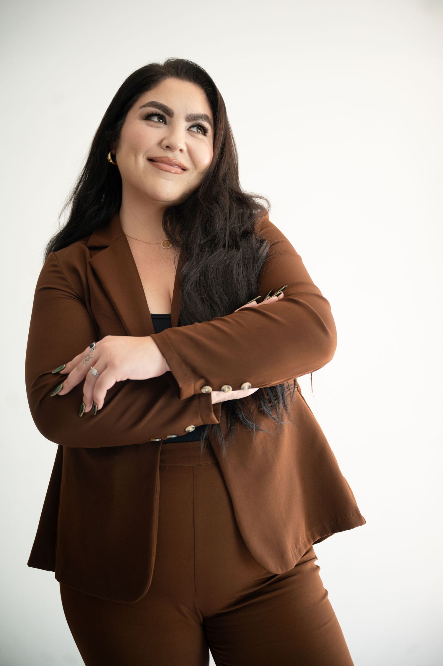 Professional portrait of someone in a brown business suit smiling and posing with arms crossed against white background.