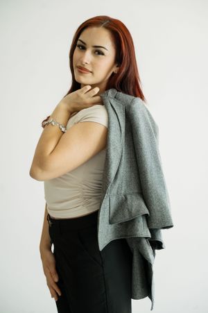 Fashion portrait with person in tan top and dark pants holding a draped gray wool coat against white background.