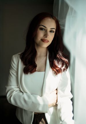 Elegant portrait featuring dark wavy hair, red lipstick, and a stylish white blazer in moody natural lighting.
