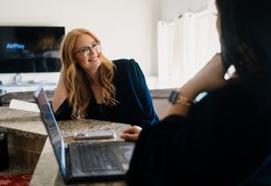 Two people having an engaging conversation at a desk with a laptop computer in a bright office setting.
