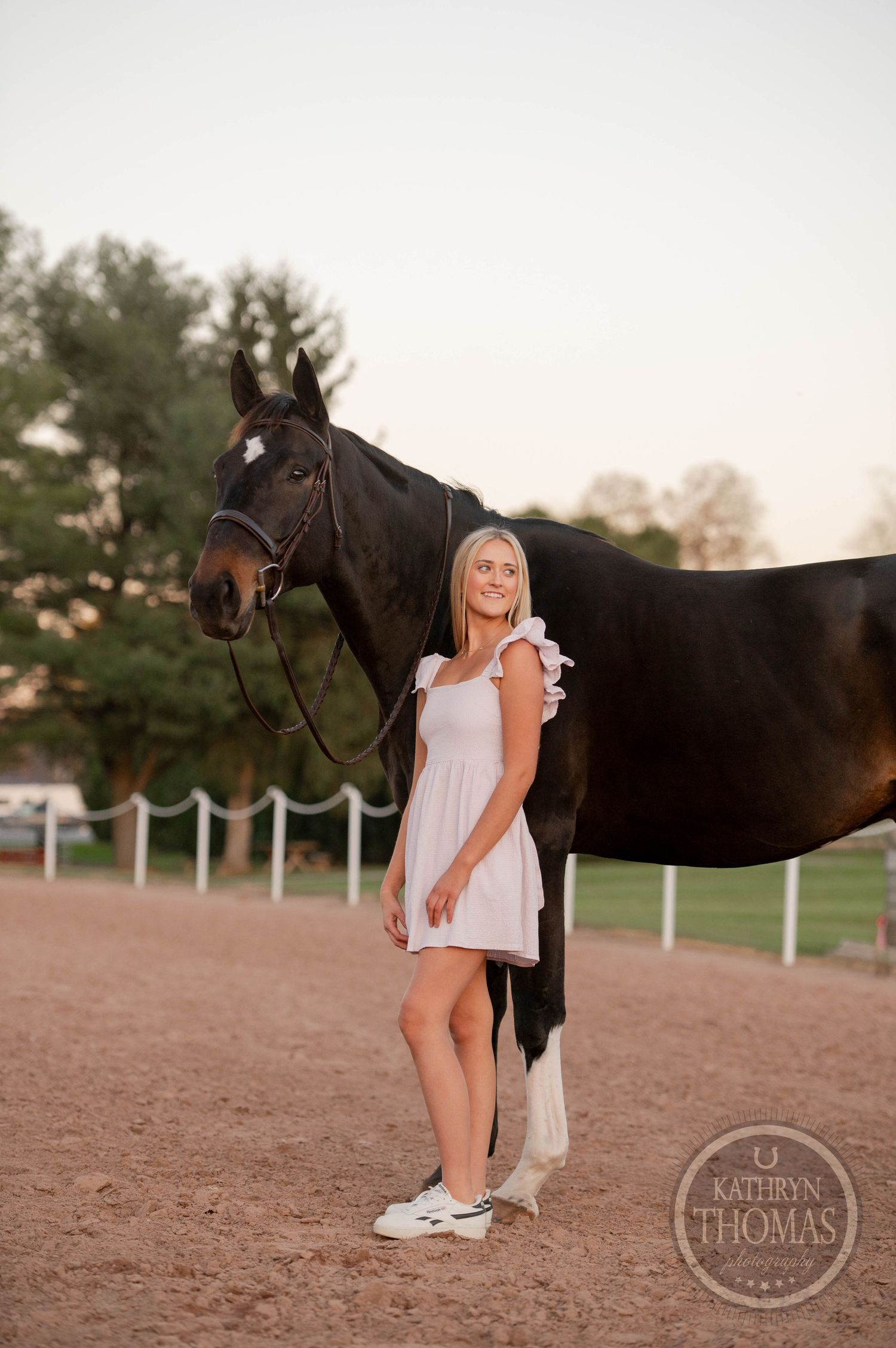 A dark horse and someone in a white dress pose together in a sandy arena during sunset.