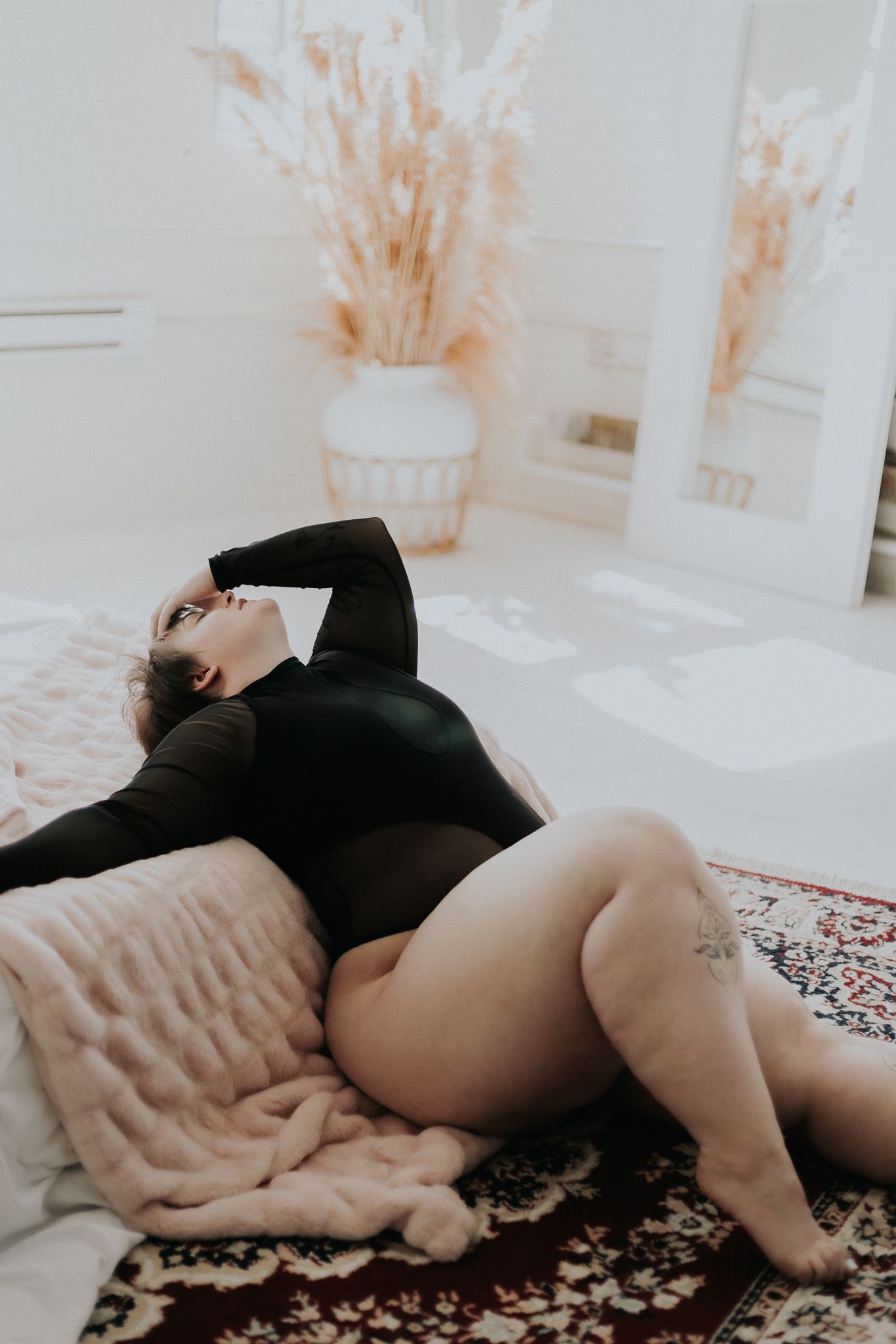 Intimate portrait in black bodysuit on pink bedding with dried pampas grass decor.