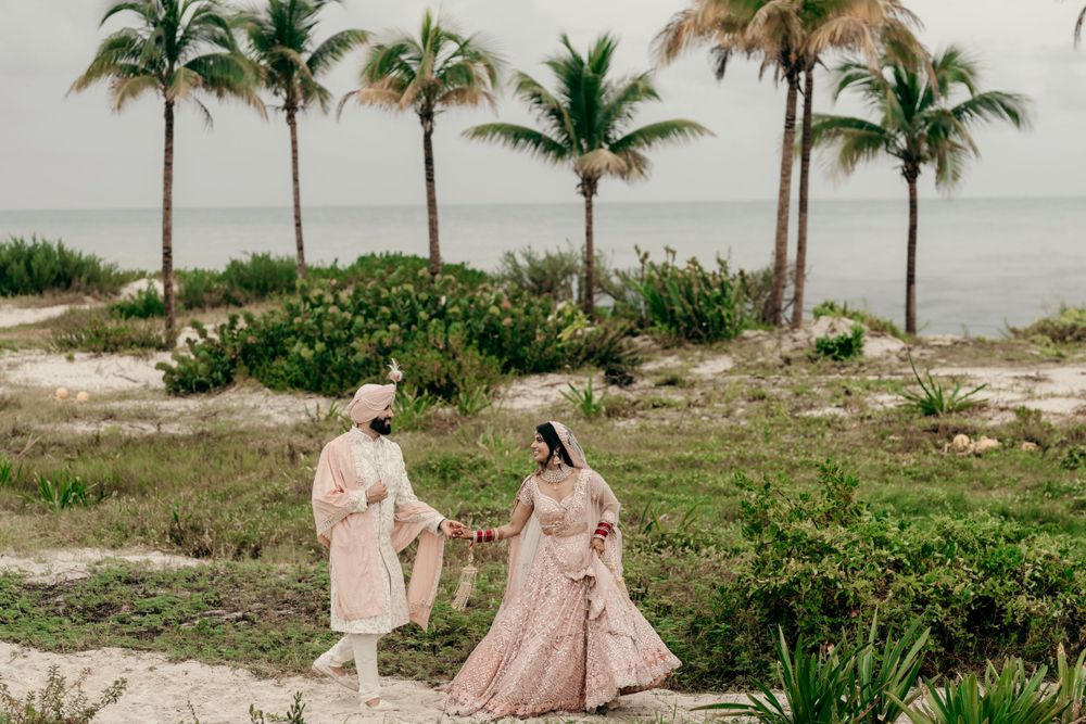 Two people in elegant attire walk along a tropical beach path lined with palm trees and coastal vegetation.