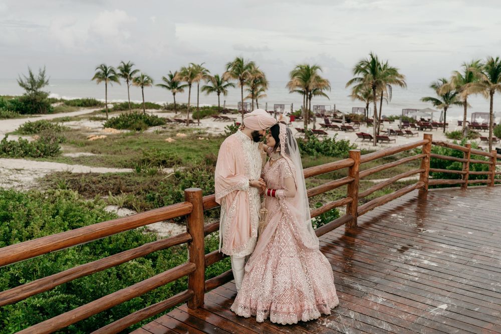 A couple in elegant pink wedding attire share an intimate moment on a wooden boardwalk by a tropical beach with palm trees.