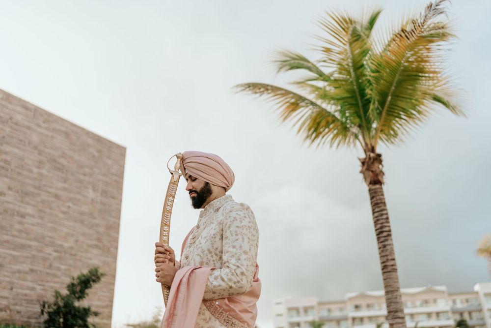 A Sikh groom in traditional attire stands beneath a palm tree against a modern building backdrop during a wedding ceremony.