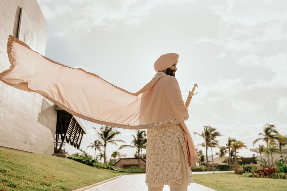 A figure in a flowing beige cape stands on a lawn with palm trees and buildings in the background.