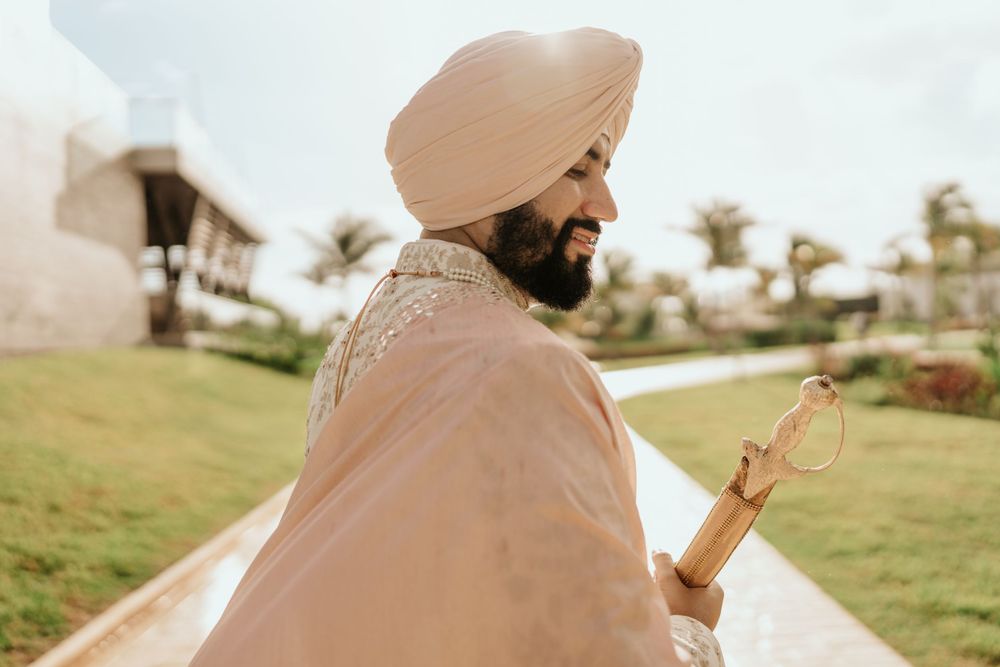A person in traditional Sikh attire with a beige turban and light-colored clothing stands in a sunlit garden setting.
