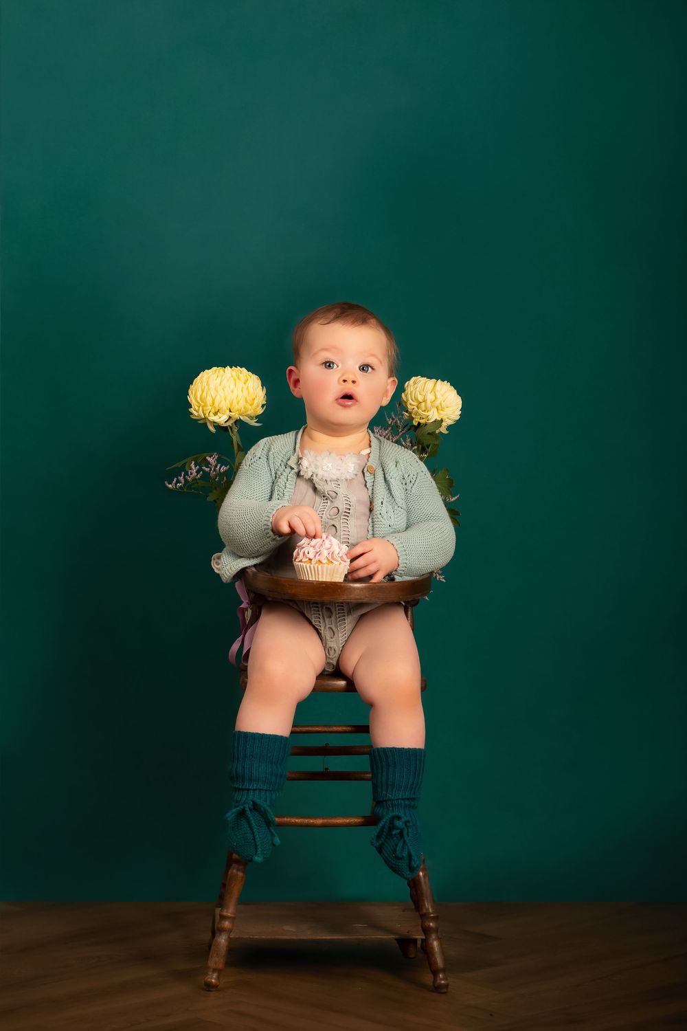 Baby holding yellow flowers sits in wooden chair against teal backdrop.