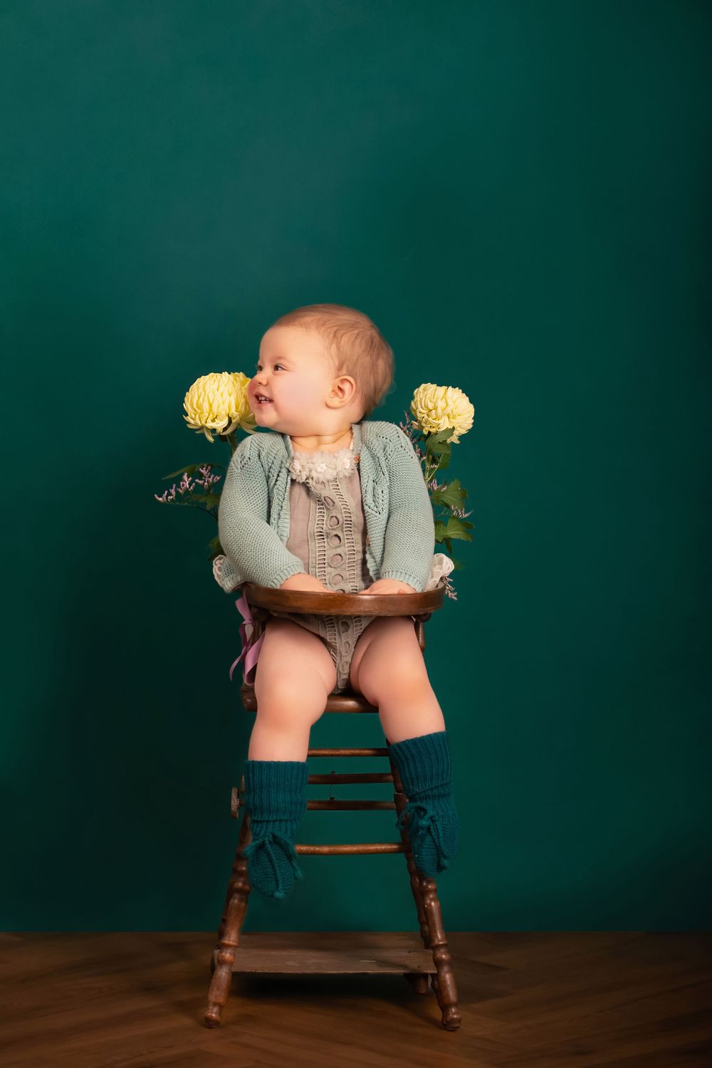 Baby wearing vintage-style outfit sits in wooden chair with yellow flowers against emerald green backdrop.