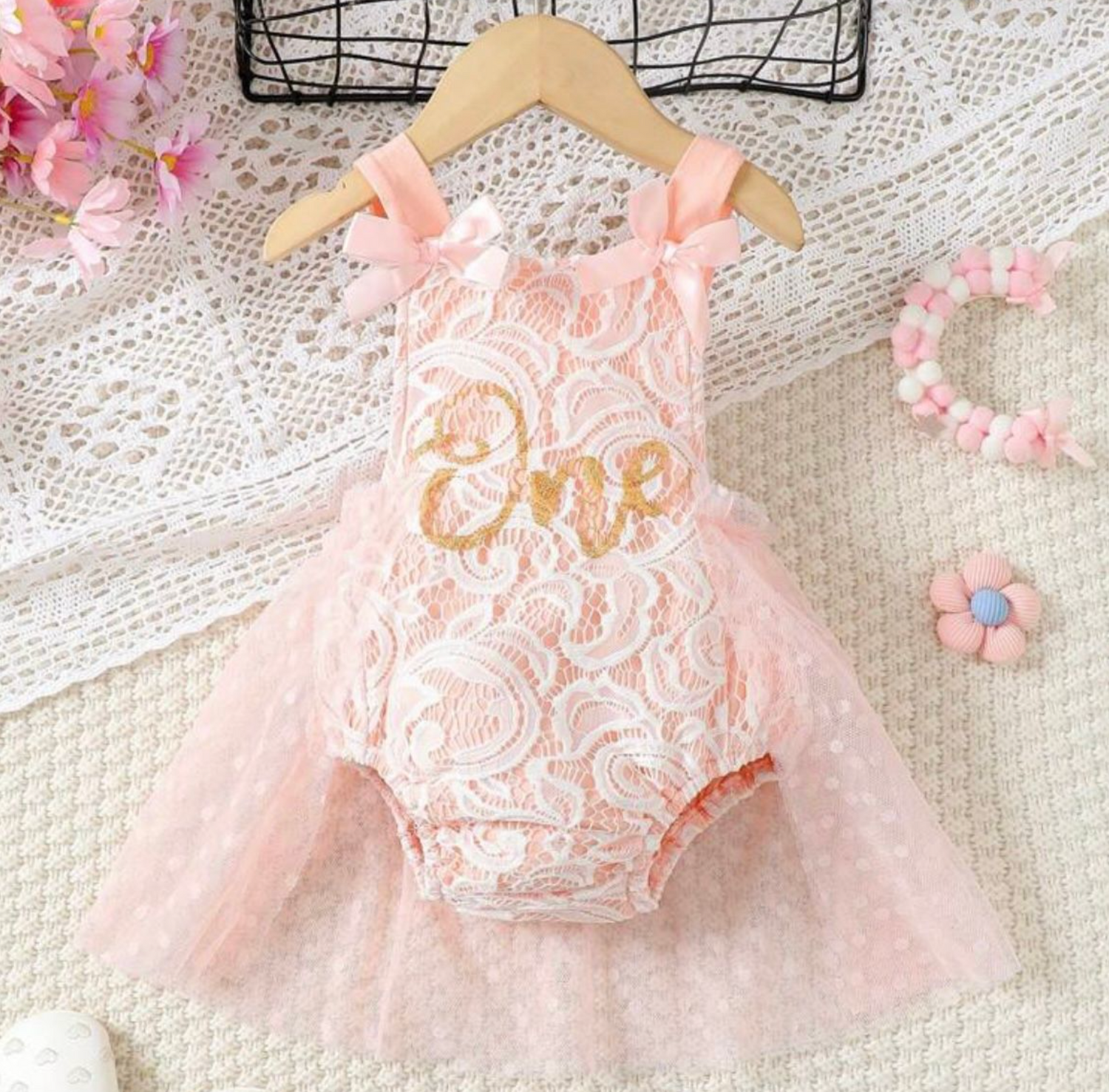 Delicate pink lace baby dress with floral details displayed on white textured background.