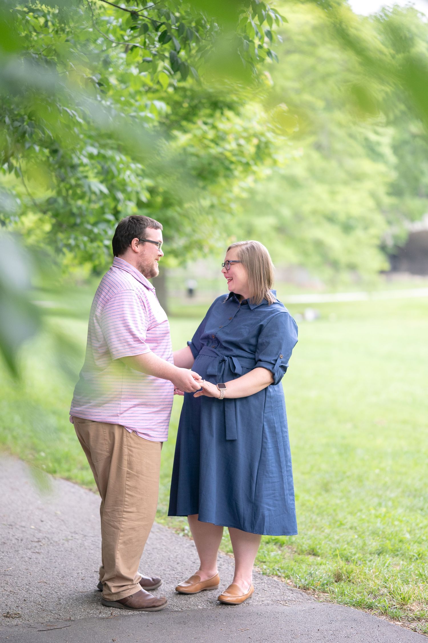 A couple stands together on a tree-lined path, sharing an intimate moment in a lush green outdoor setting.