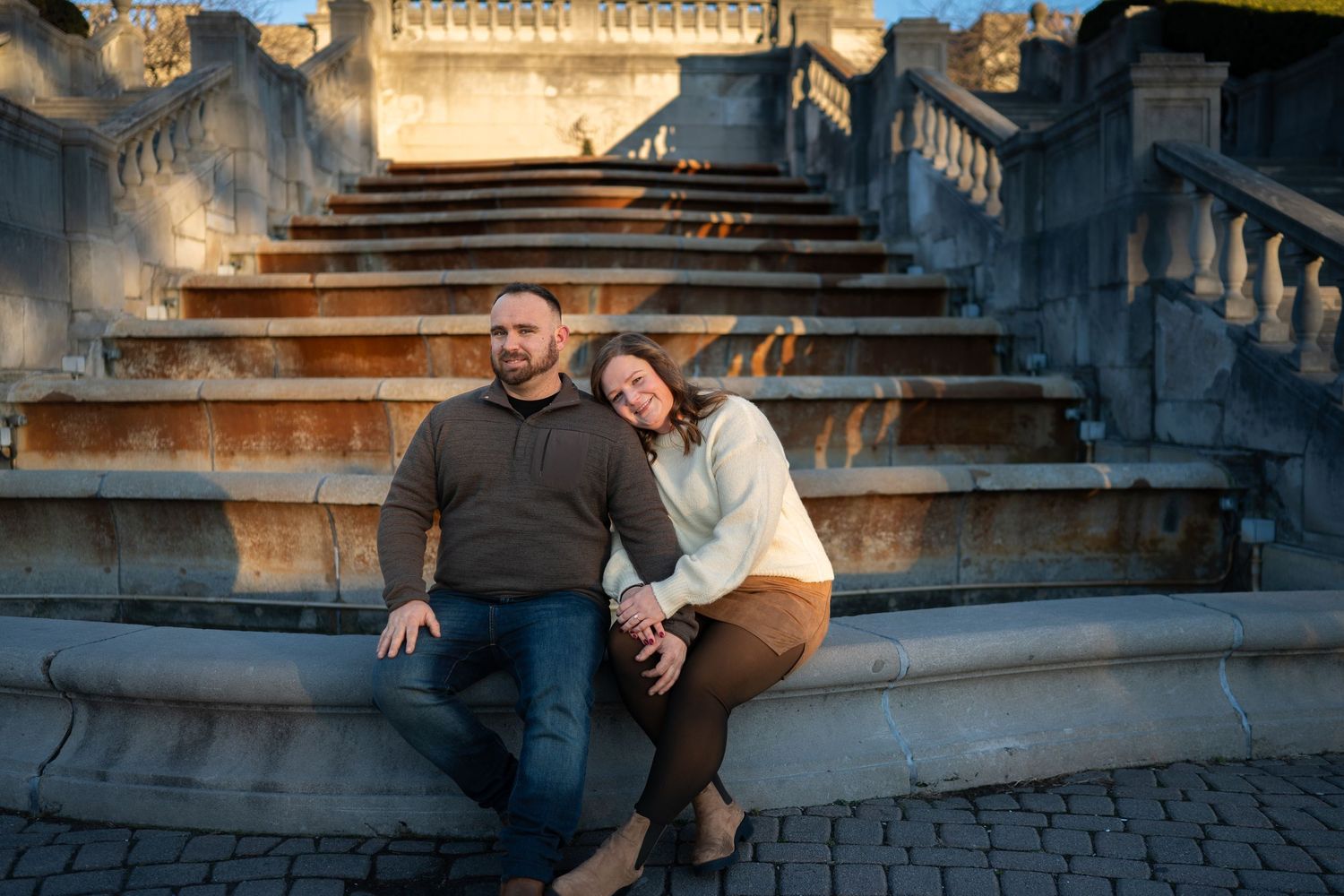 A couple sits together on stone steps during golden hour with warm sunlight and architectural details in the background.