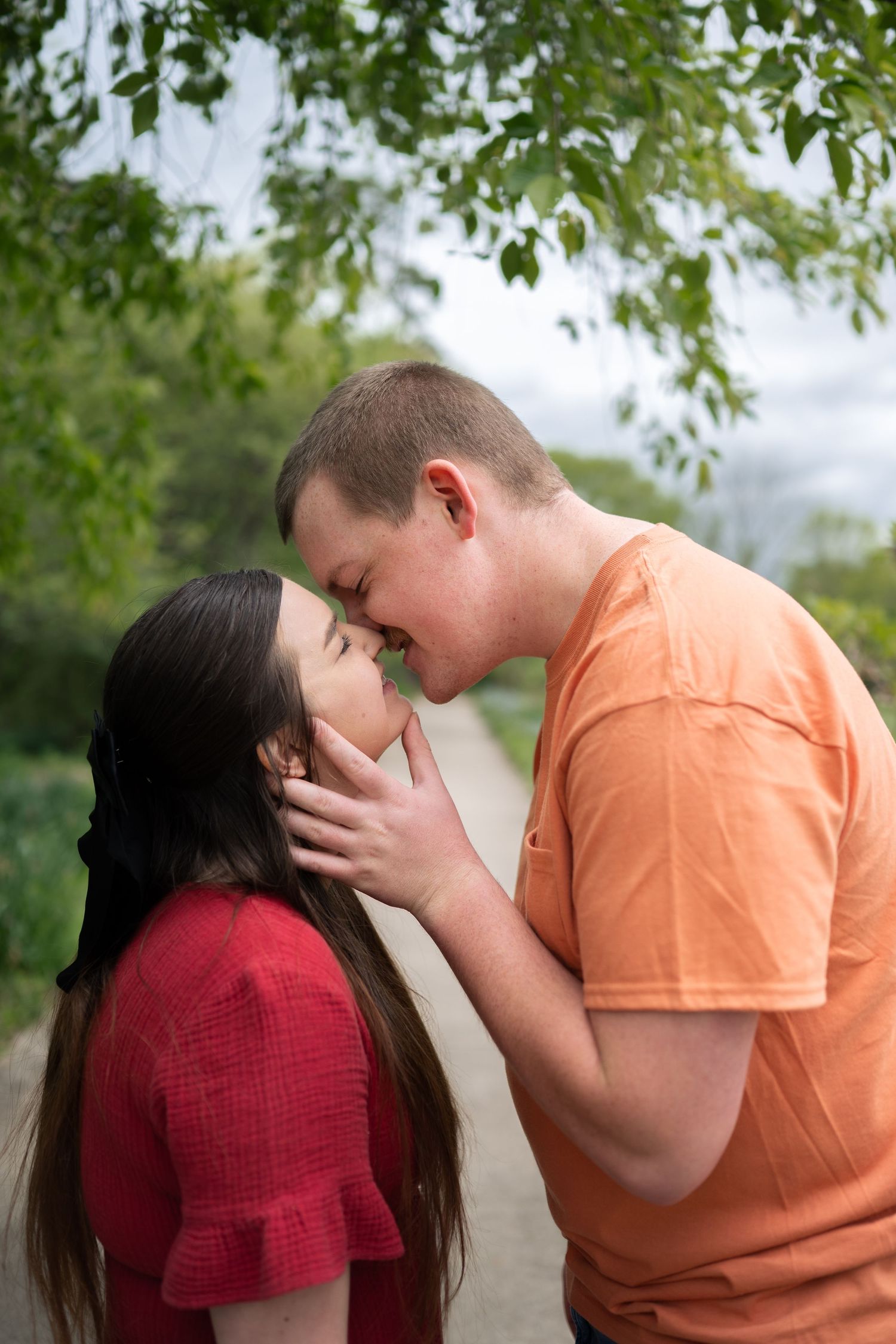 A young couple shares a romantic moment, nearly kissing outdoors under green tree branches.