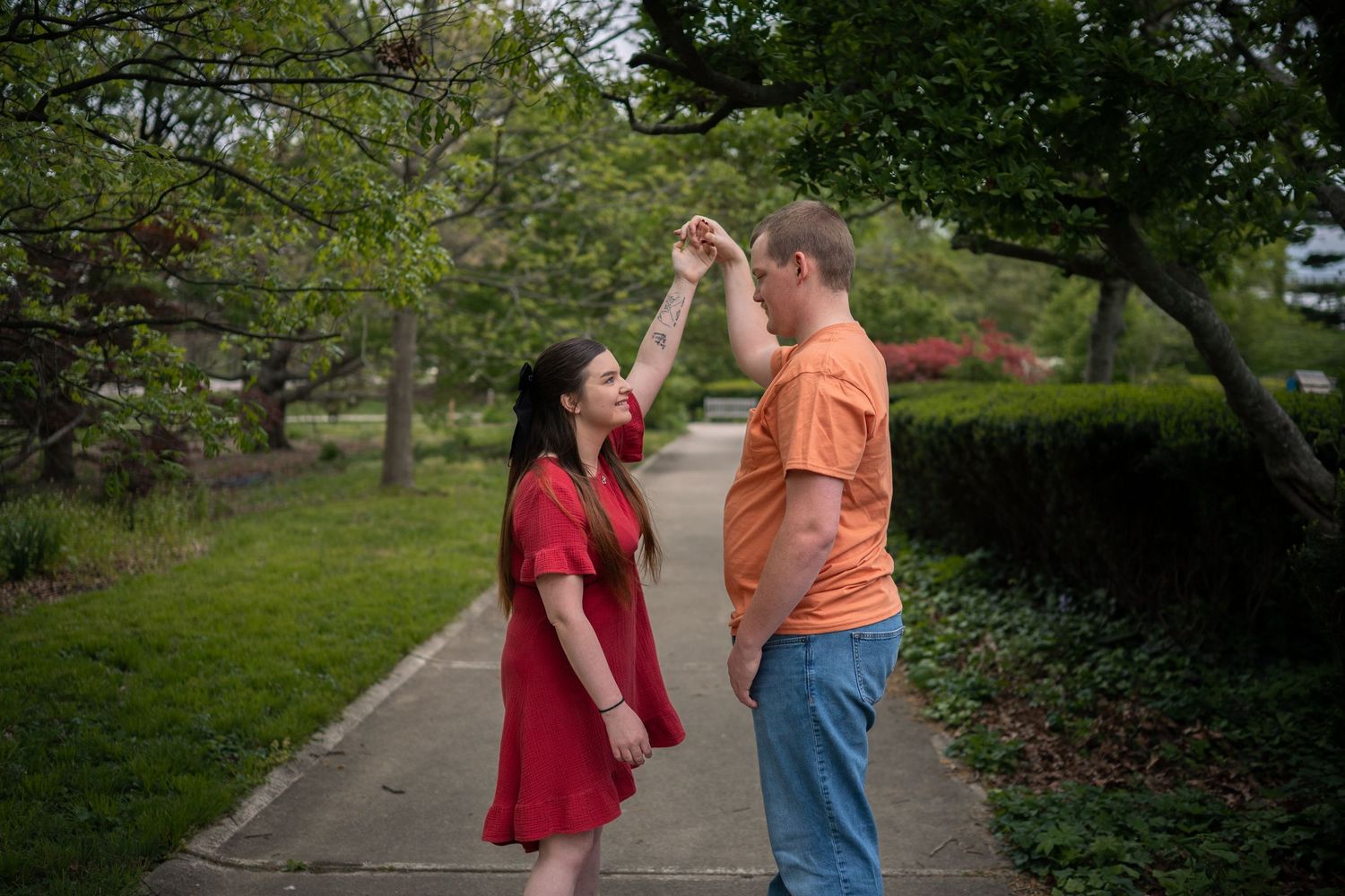 Couple dancing on a park path, woman in red dress, man in orange shirt, surrounded by lush green trees.