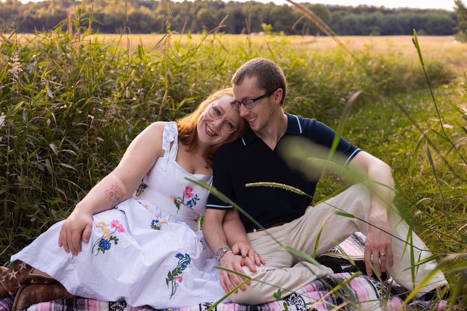 Couple sitting together in a lush green field, woman in white floral dress leaning on man in navy polo at sunset.