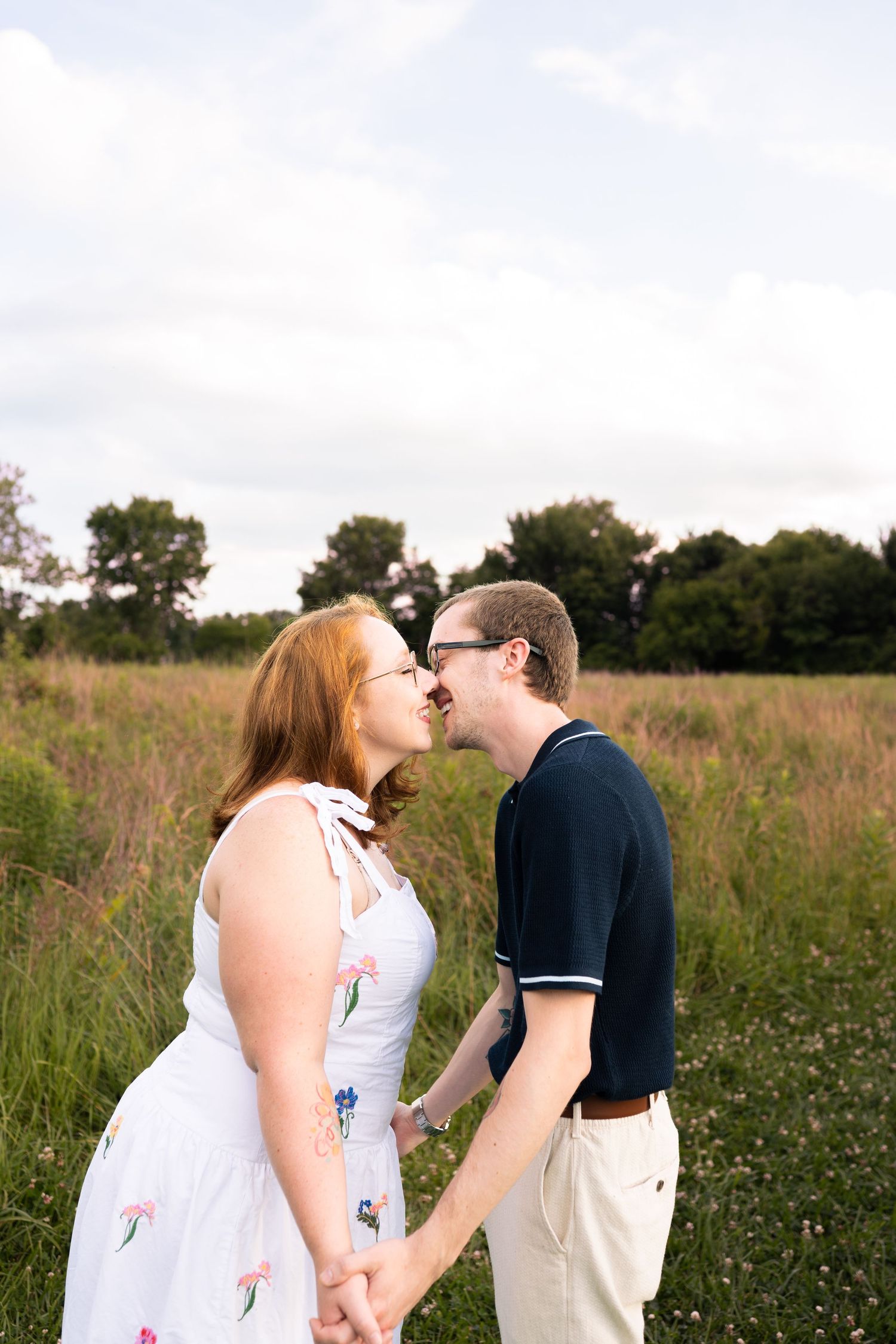 Couple holding hands and sharing a kiss in a lush green meadow, woman in floral dress, man in navy polo shirt.