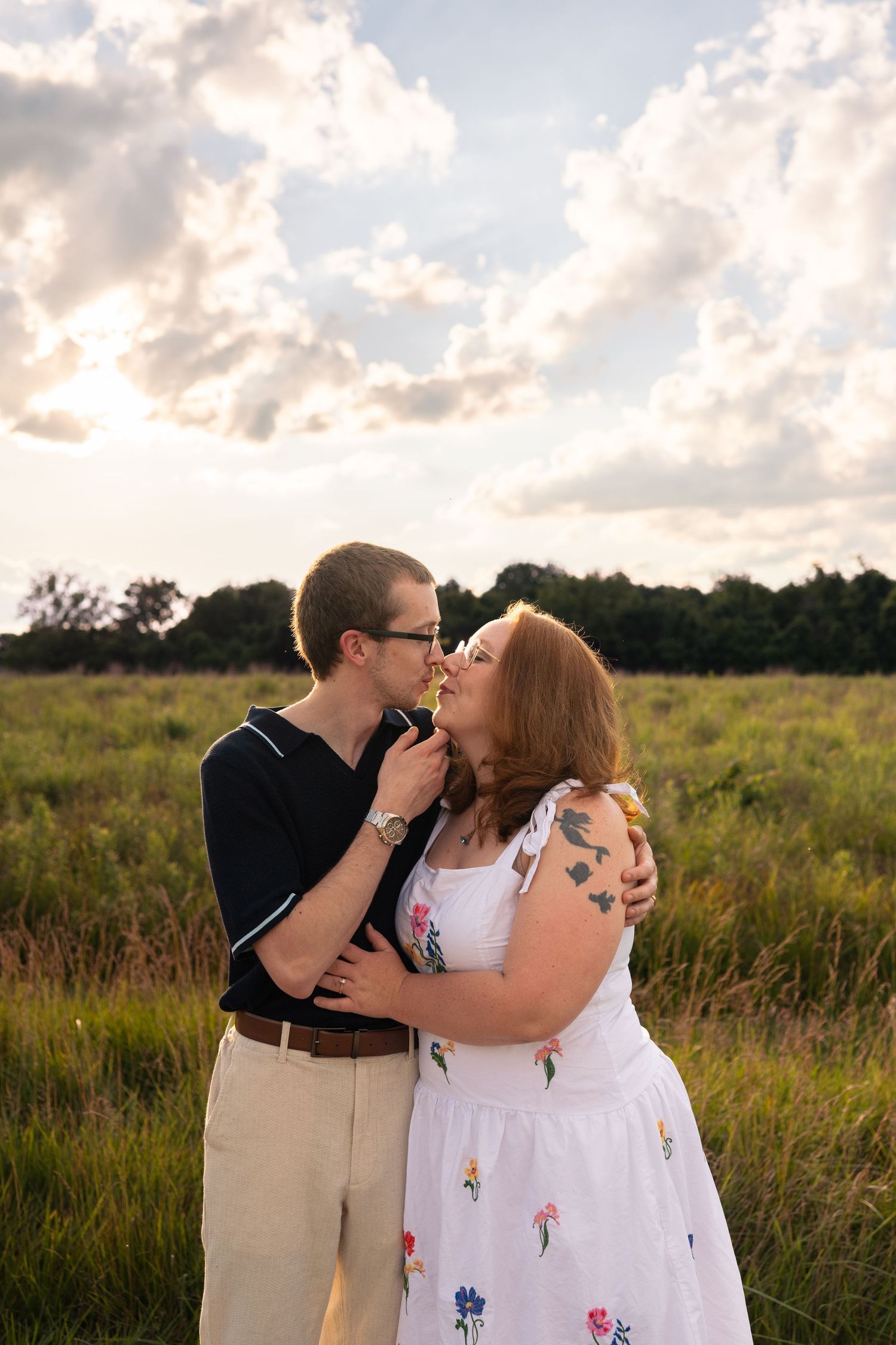 Couple sharing a romantic moment in a green meadow, woman in floral dress and man in black shirt, golden hour sky.