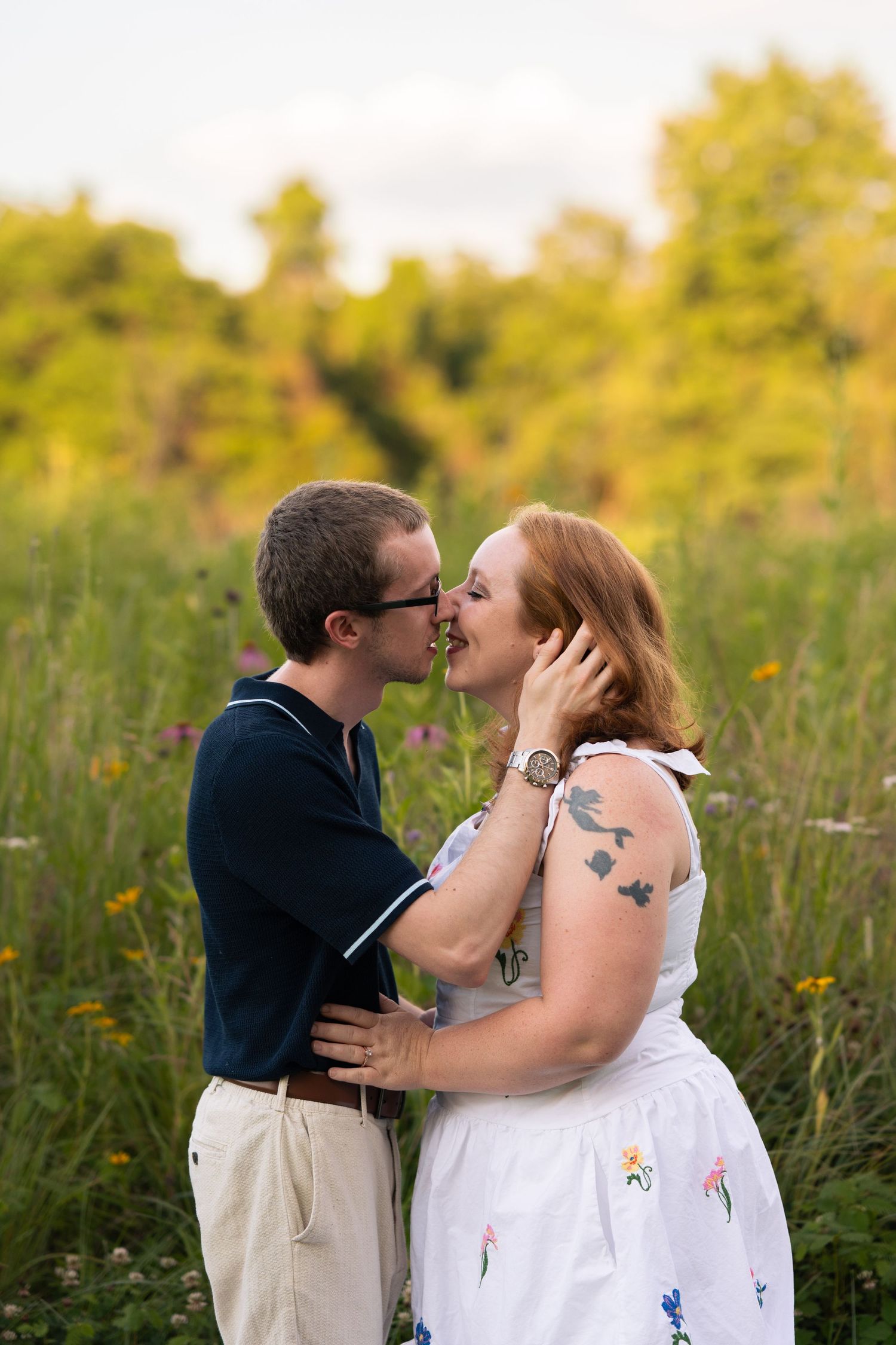 A couple shares a romantic moment in a wildflower meadow, the woman in a white dress with tattoos, the man in a navy polo.
