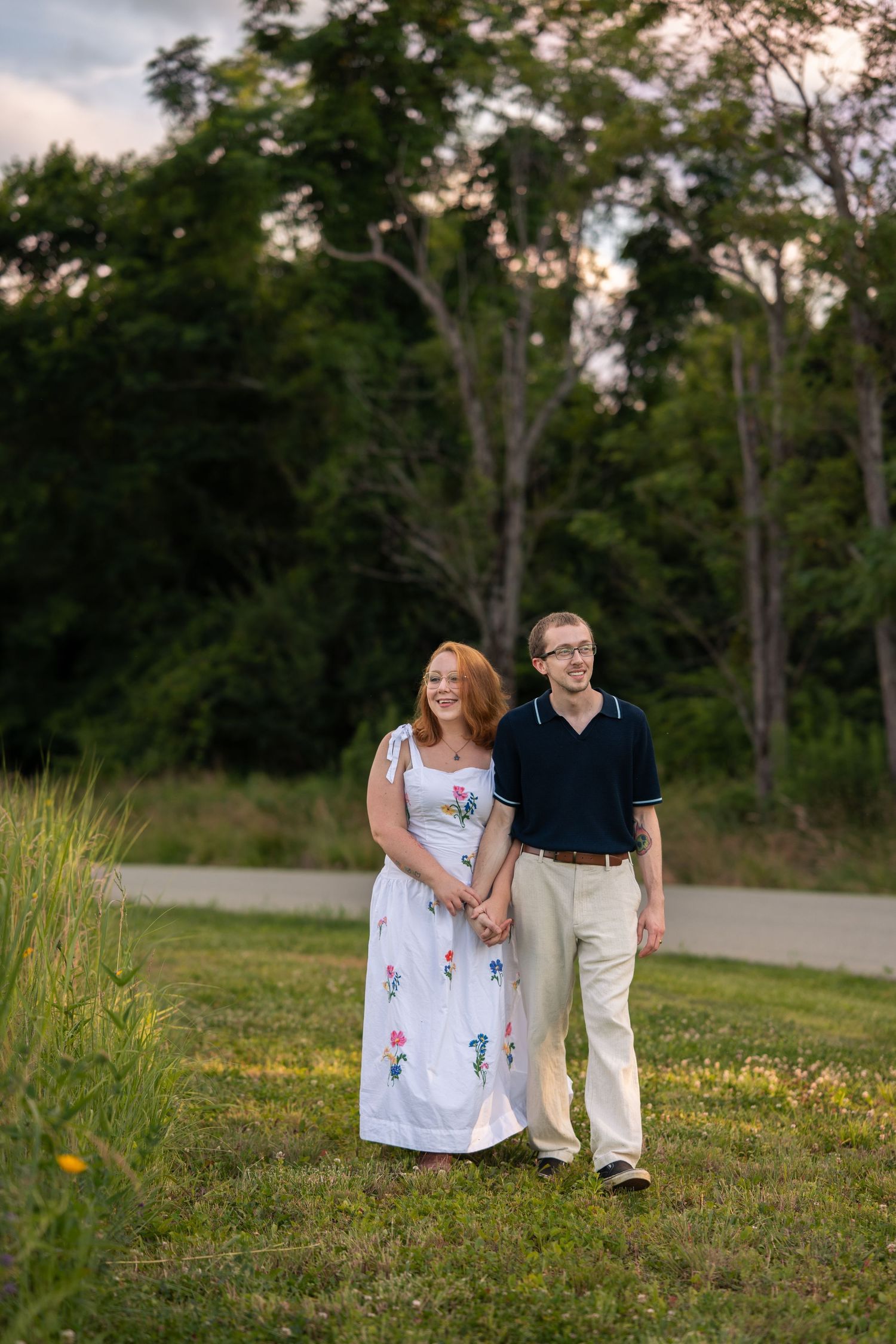 A couple holds hands smiling in a green field, woman in floral dress, man in navy polo, trees in background.