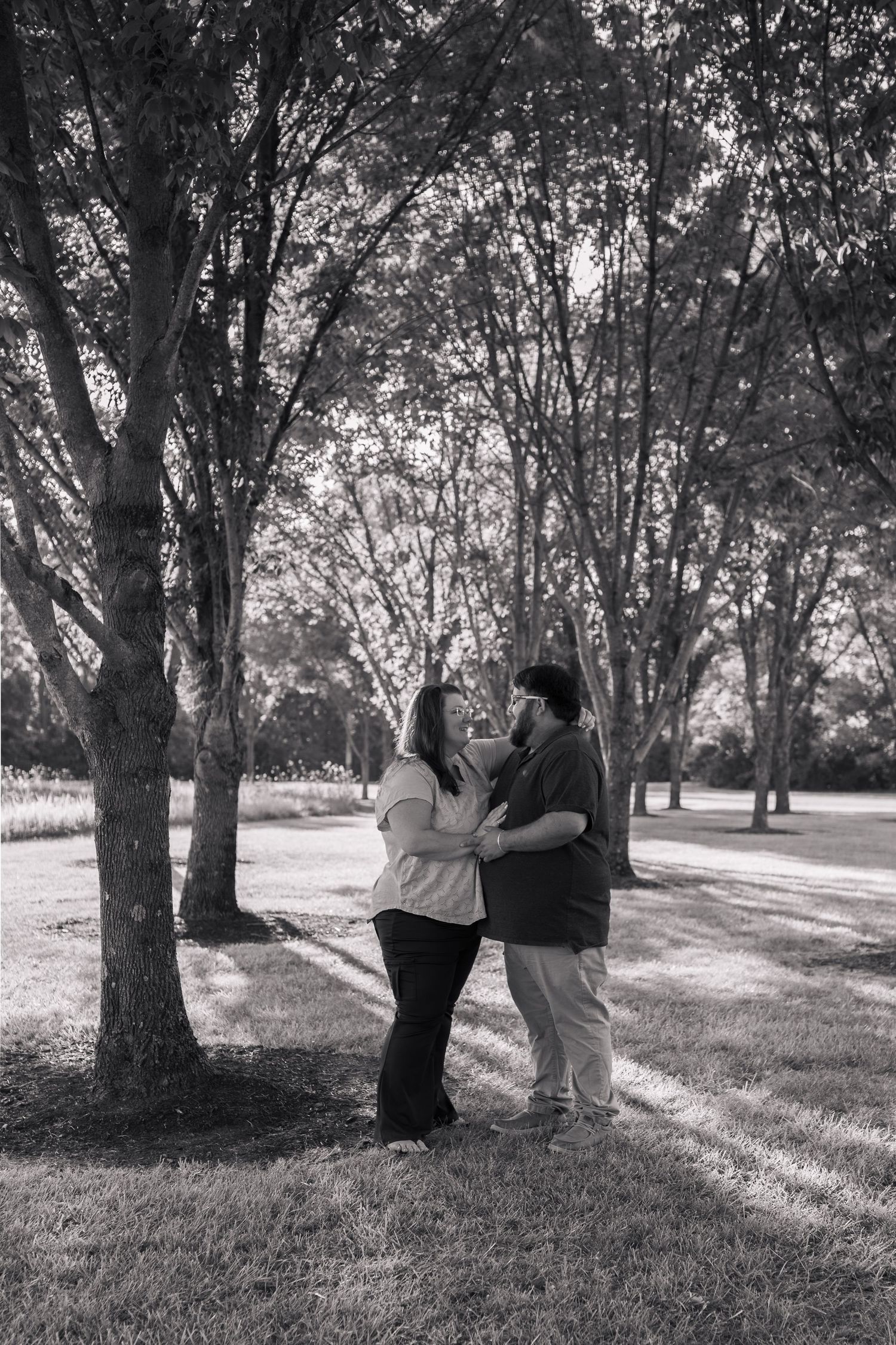 Black and white photo of a couple holding hands and gazing at each other in a tree-lined park.