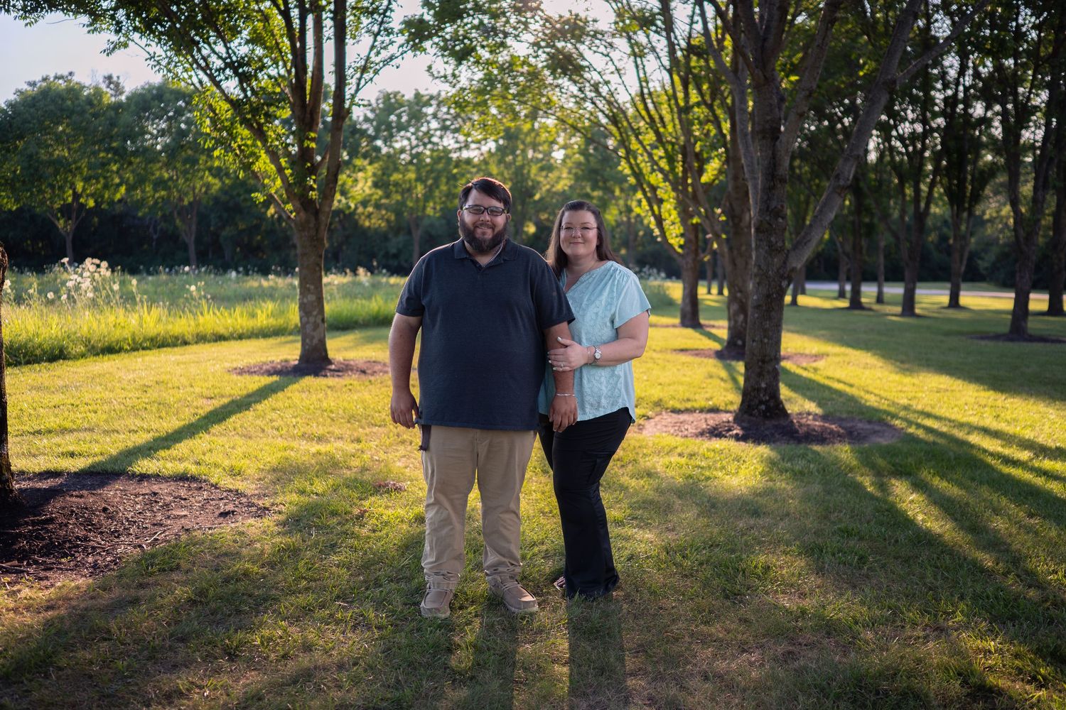 A couple smiles together in a lush green park surrounded by tall trees during golden hour sunlight.